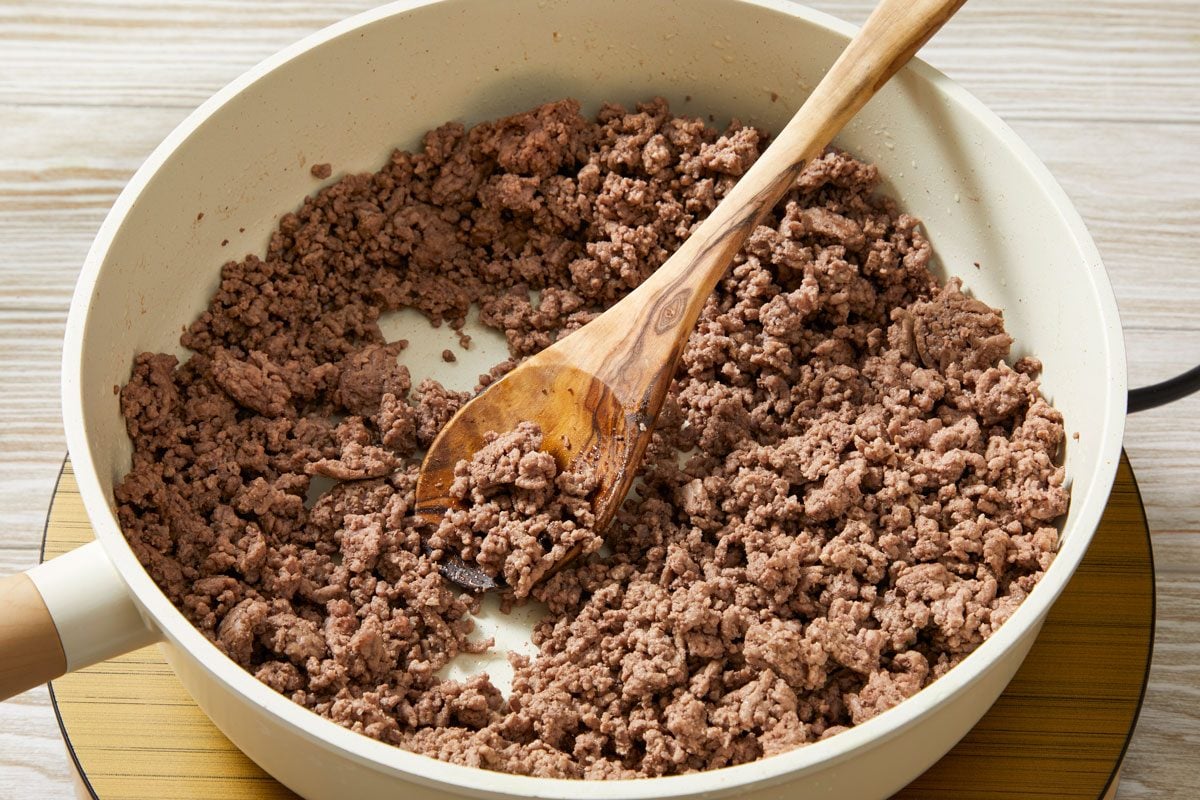 Close shot of a large skillet cook beef over medium heat until no longer pink; drain; wodoen spoon; induction; cream-colored wooden surface;
