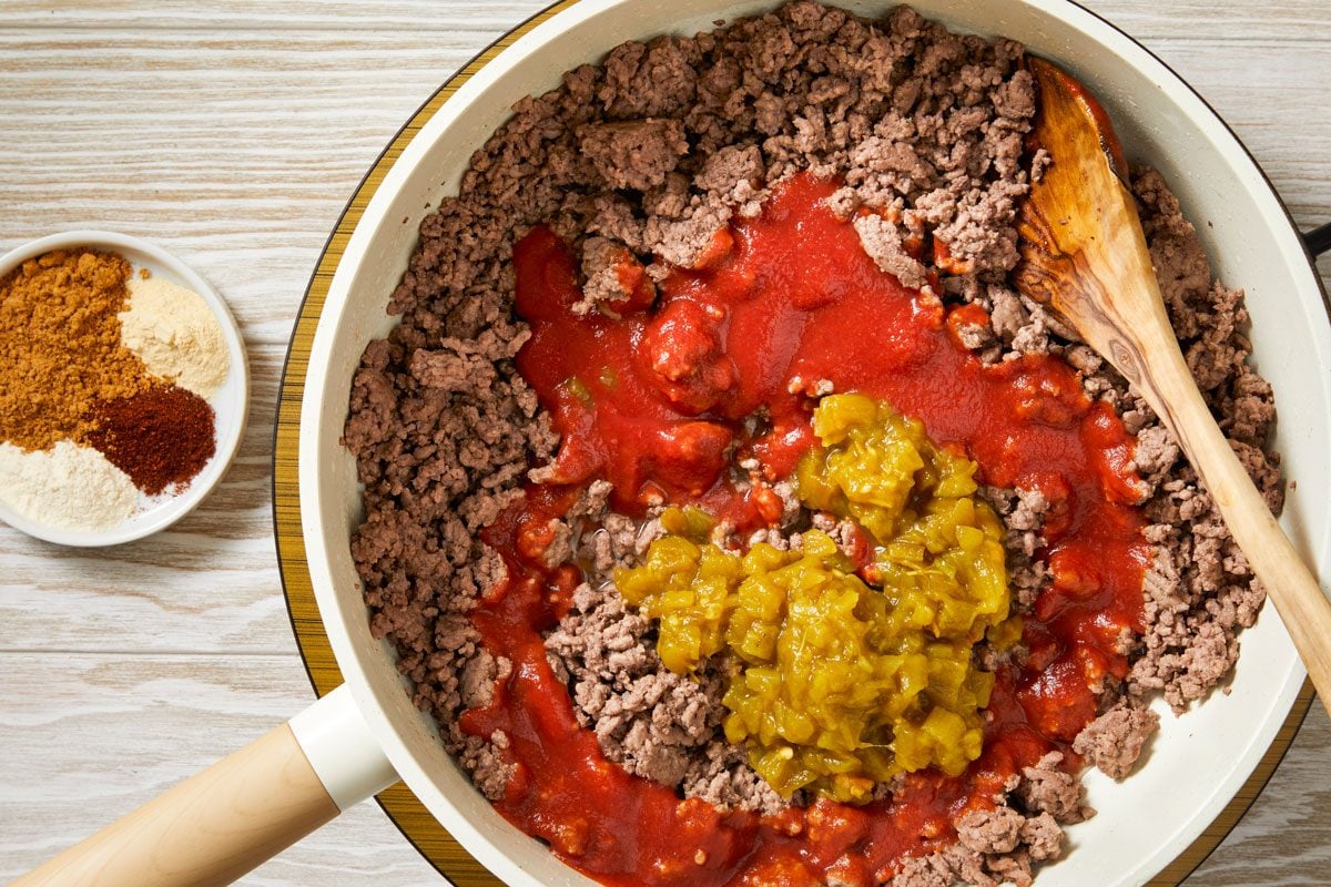 Overhead shot of stir in tomato sauce; water; green chiles; taco seasoning; onion powder; chili powder and garlic powder; Bring to a boil; Reduce heat; simmer uncovered 5 minutes; wooden spoon; induction; cream-colored wooden surface;