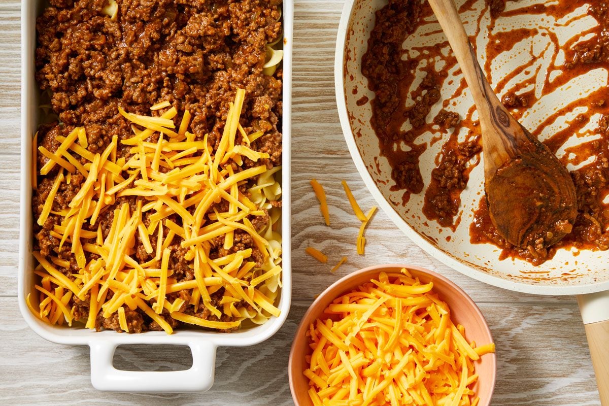 Overhead shot of spread beef mixture over noodles layer; sprinkle top with cheese; Bake uncovered until cheese is melted 10-15 minutes; Let stand 10 minutes; cheese bowl; wodoen spoon; cream-colored wooden surface;