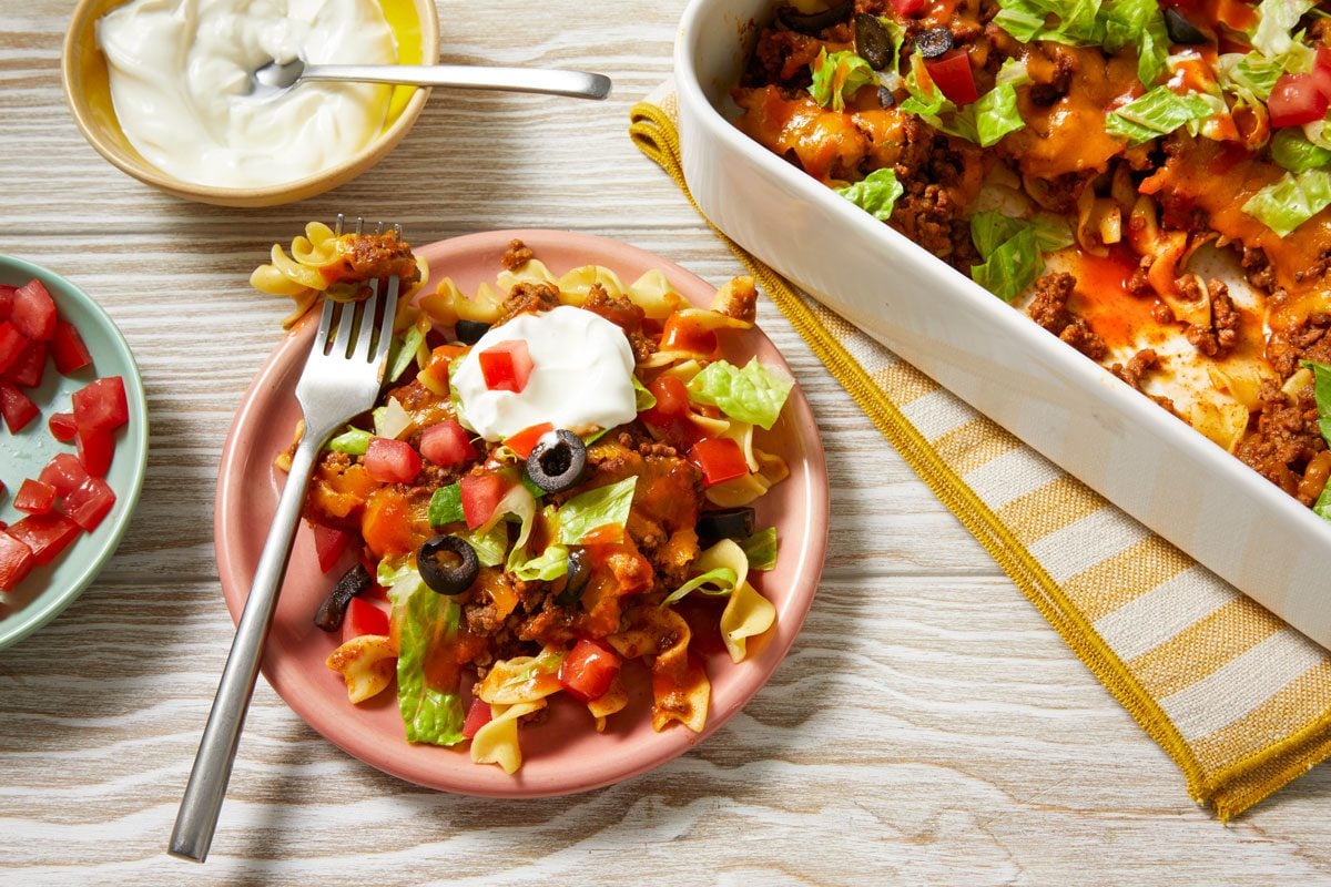 Overhead shot of Taco Noodle Bake in a baking pan; Top with lettuce; tomatoes; olives and taco sauce; a portion served on a small plate garnished with sour cream; a fork and a yellow and white linen napkin; set on a cream-colored wooden surface;