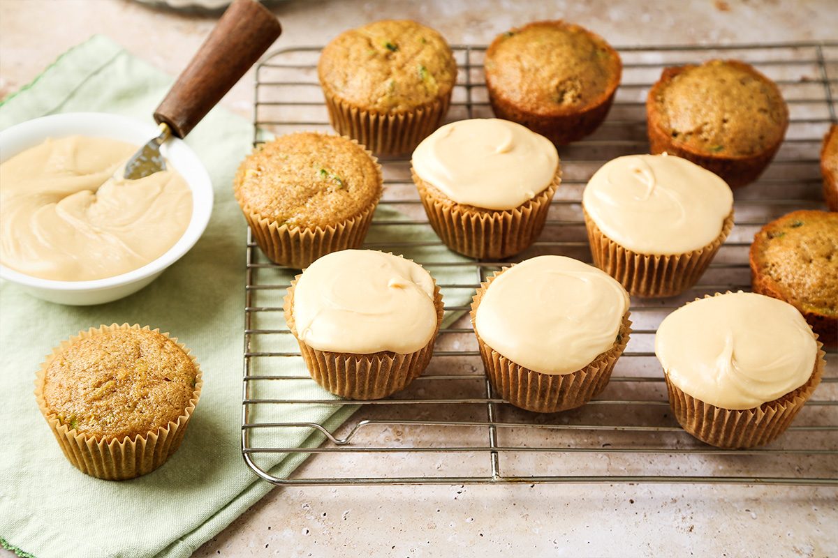 A cooling rack holds several cupcakes, some frosted with light brown icing and some plain. A bowl of icing with a spatula rests on a green cloth beside the cupcakes.