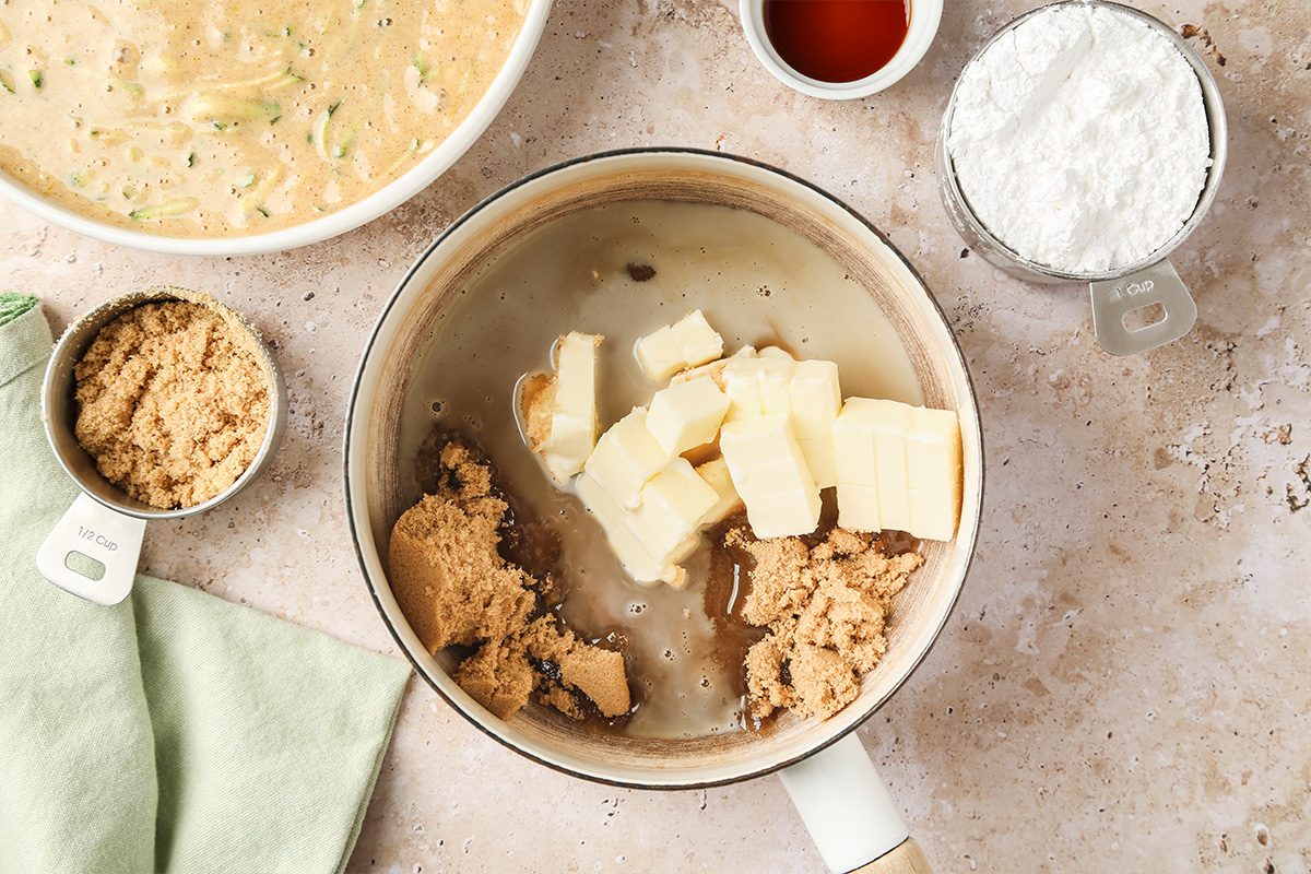 A saucepan with butter, brown sugar, and liquid ingredients sits on a counter beside a bowl of batter, a measuring cup of powdered sugar, and a small cup of vanilla extract. A green cloth is nearby.