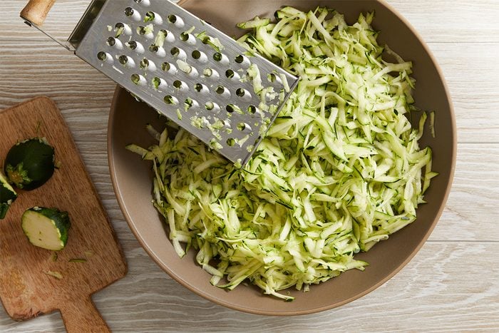 A bowl filled with freshly grated zucchini beside a metal grater, with zucchini ends placed on a wooden cutting board nearby, all set on a light wood surface.