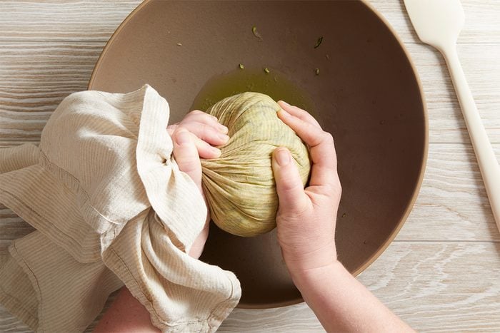 Two hands squeeze shredded zucchini wrapped in a cloth over a brown bowl, extracting liquid. A spatula rests on a light wooden surface beside the bowl.