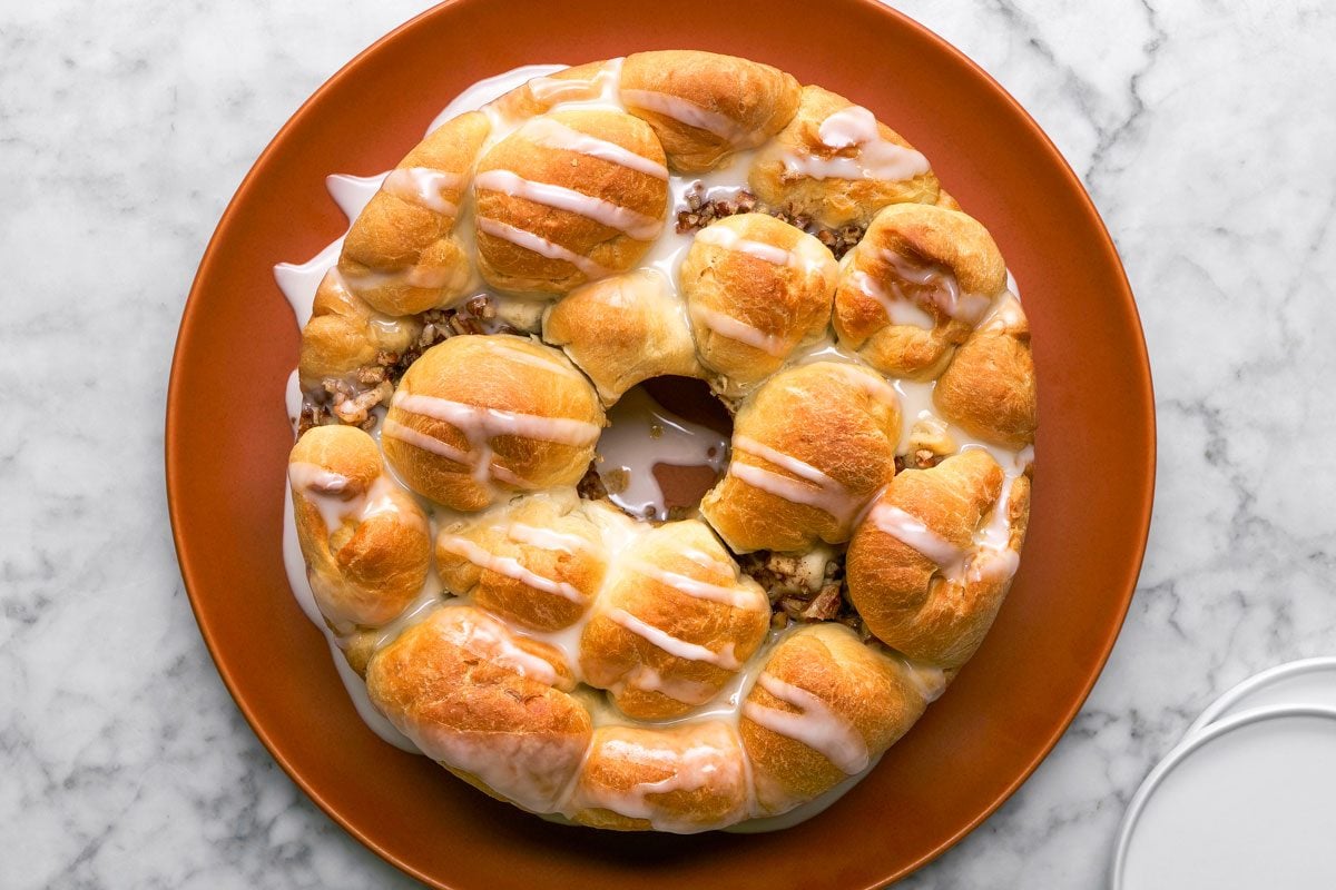 overhead shot of a round pull apart apple bread ring with golden brown rolls, drizzled with white icing, sits on an orange plate atop a marble surface