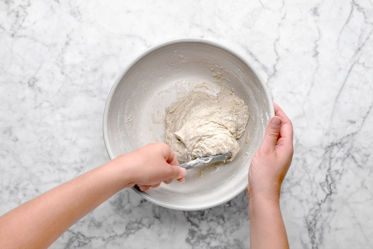 overhead shot of a person’s hands mix bread dough in a metal bowl with a spatula on a white marble surface