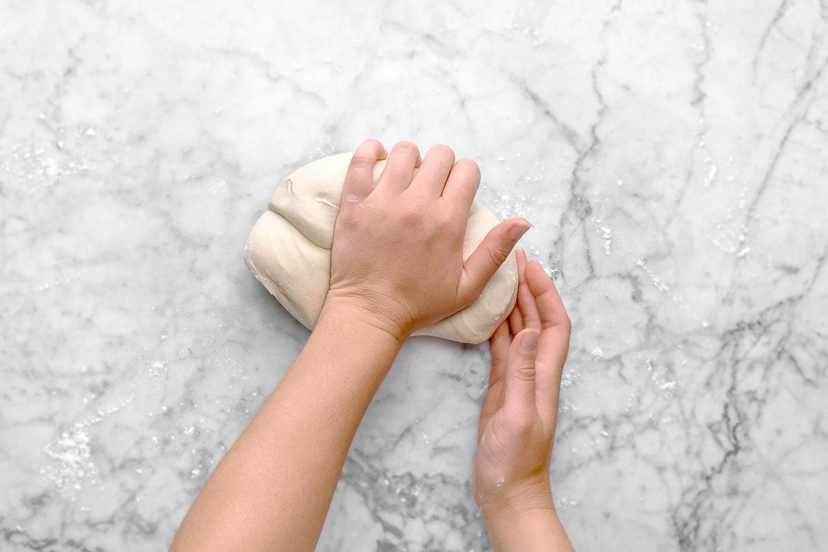 overhead shot of of two hands kneading a ball of dough on a lightly floured marble surface