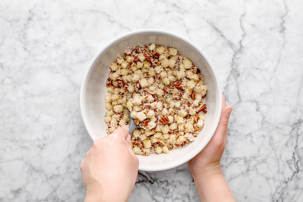 overhead shot of a person’s hands mix a crumbly mixture of flour, sugar, and pecans in a large white bowl on a marble countertop