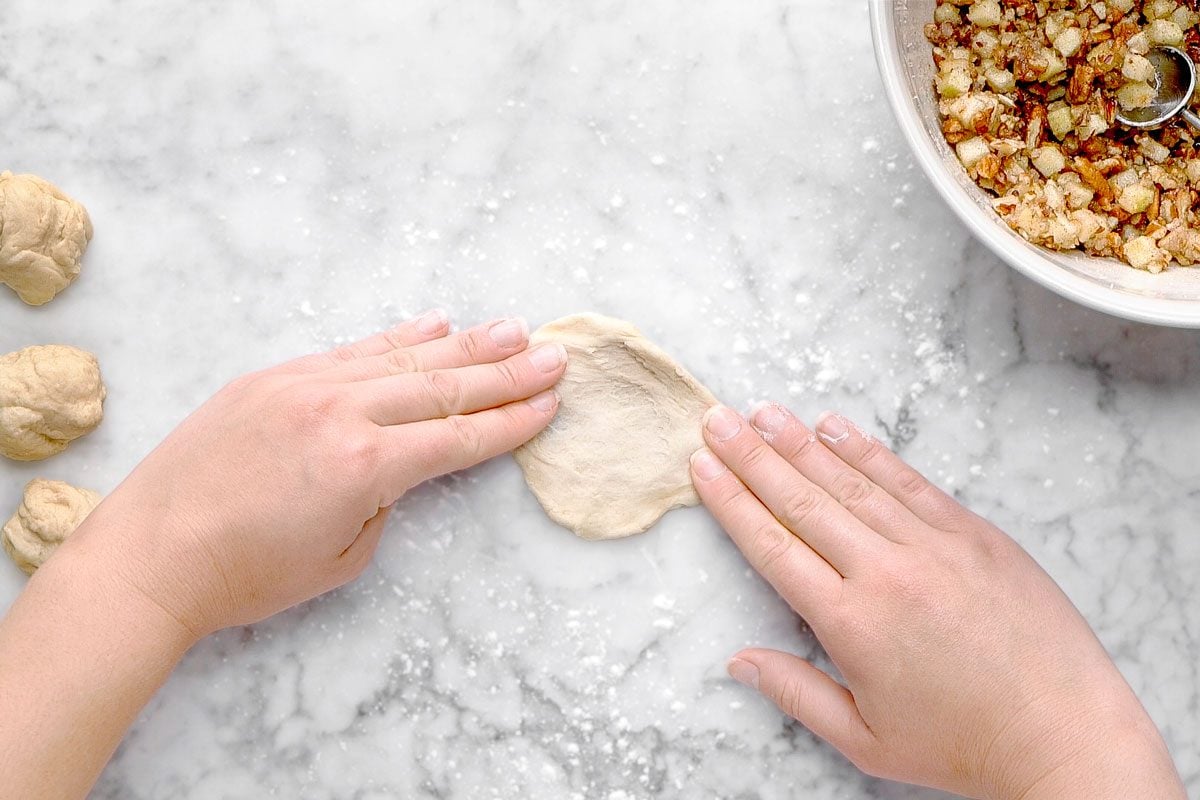 overhead shot of Hands flatten a piece of dough on a floured marble surface, with small dough balls on the left and a bowl of chopped filling with a spoon on the right