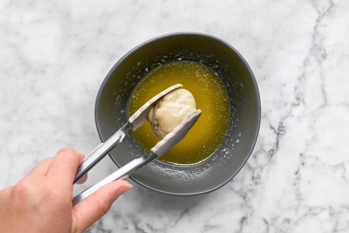 overhead shot of a hand uses metal tongs to dip a dough ball into a bowl of melted butter on a marble countertop