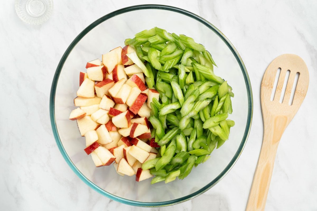 Overhead shot of a glass bowl with celery and apples tossed in lemon juice; a flat wooden spoon nearby on a marble surface;