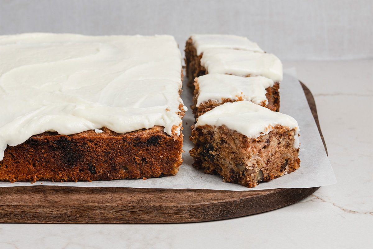 A rectangular frosted cake sits on a wooden board, with three square pieces cut and separated from the main cake, showing a moist, nutty interior. The cake is topped with a thick layer of white icing.