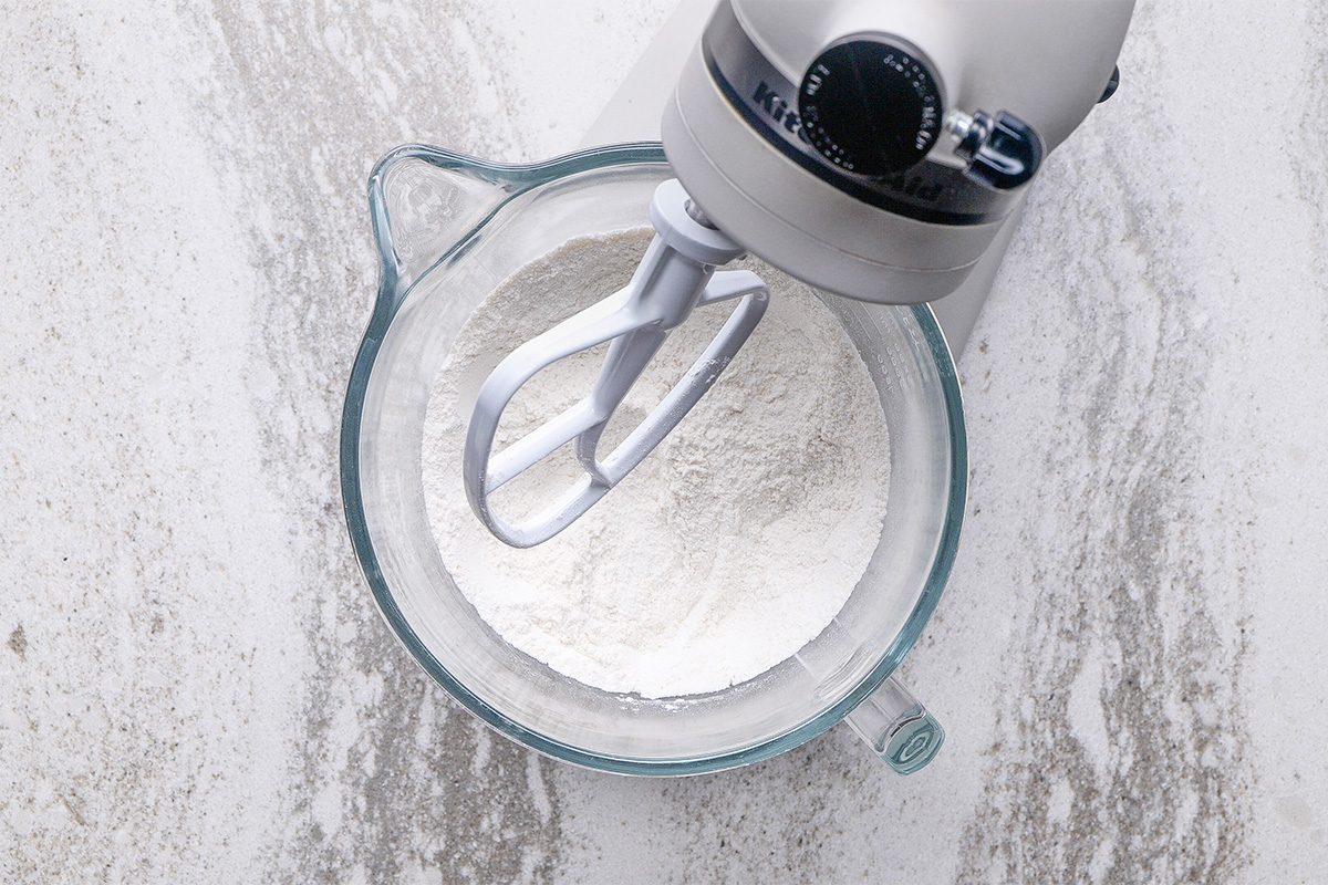 A stand mixer with a paddle attachment is mixing flour in a clear glass bowl on a light, textured countertop.