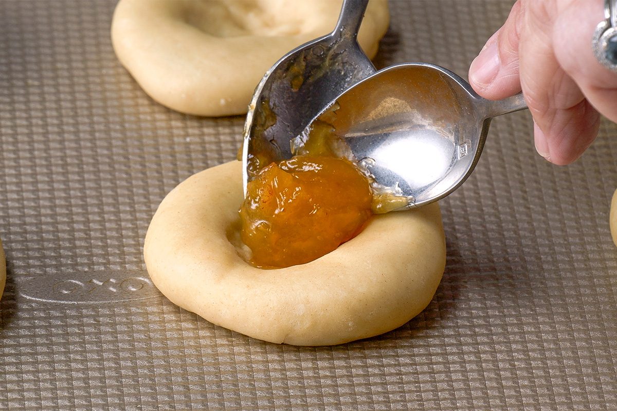 A hand uses two spoons to fill a thumbprint cookie with orange jam on a baking sheet.