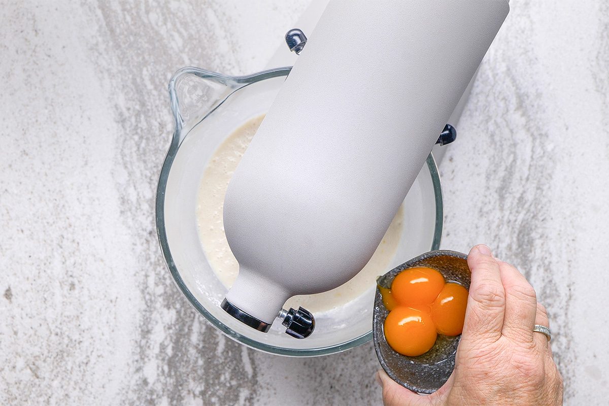 A hand holds a small bowl with three egg yolks above a stand mixer with a glass bowl containing batter on a light-colored countertop.