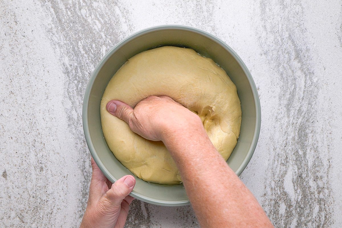 A person’s hand presses down into a bowl of risen dough, deflating it, on a light, marbled countertop.