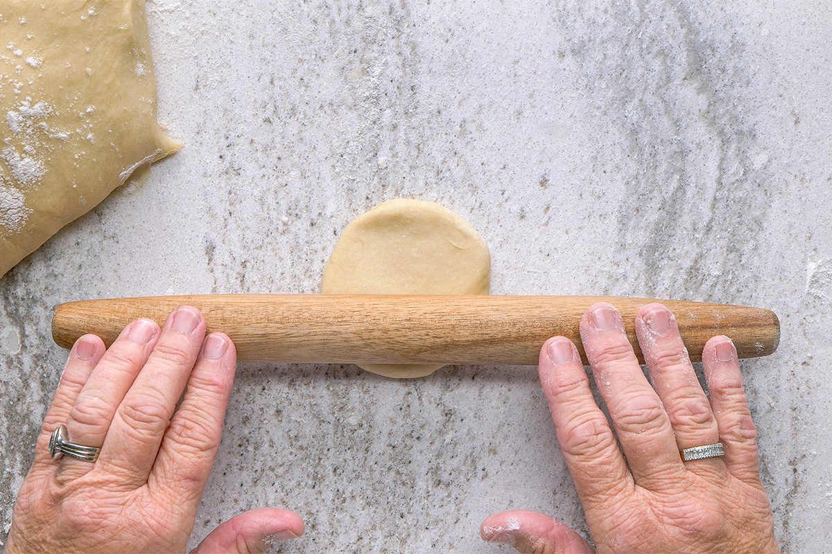 A person uses a wooden rolling pin to flatten a small round piece of dough on a lightly floured surface, with dough and flour visible nearby.