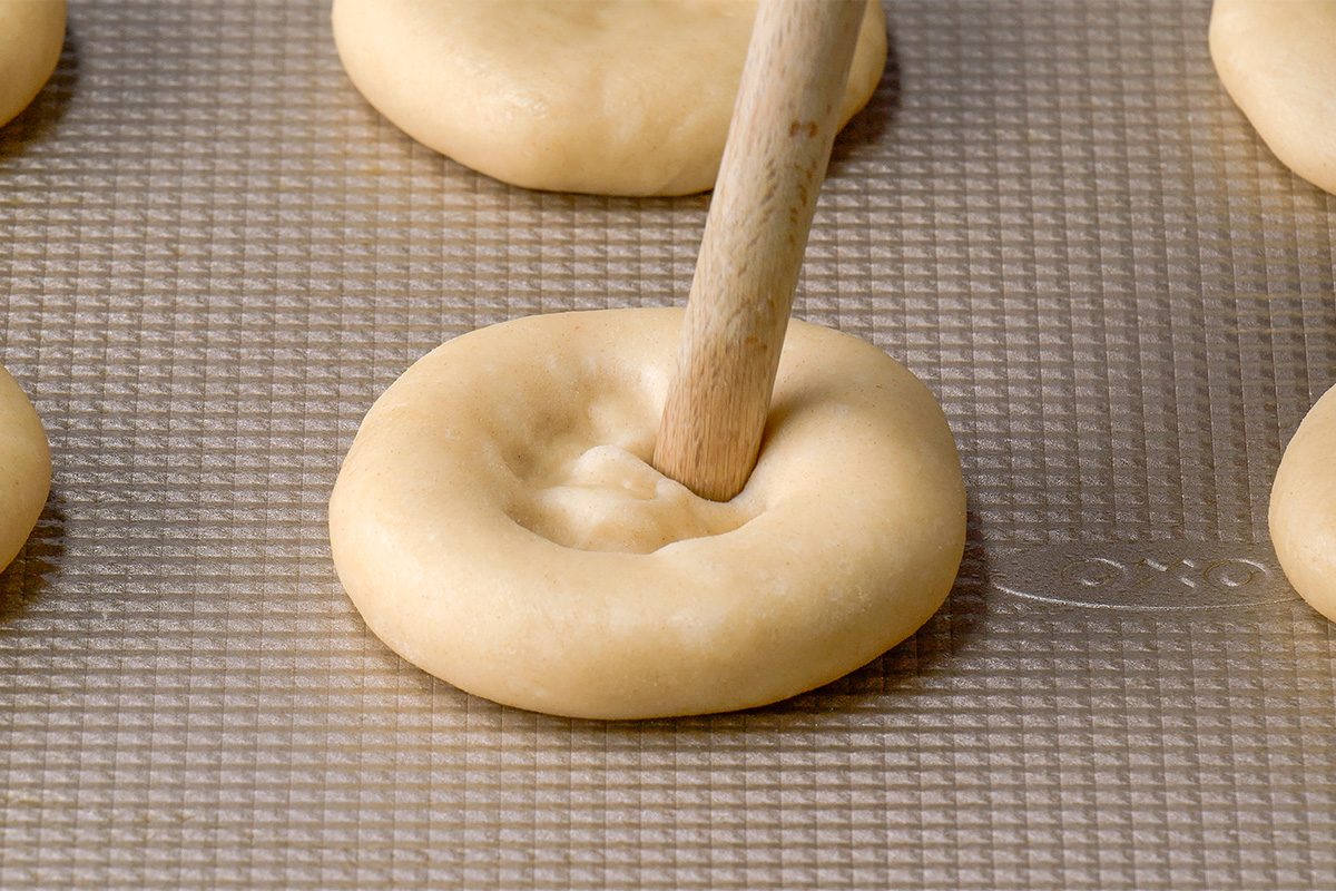 A wooden spoon handle presses a small indentation into the center of round, unbaked cookie dough on a textured baking sheet.