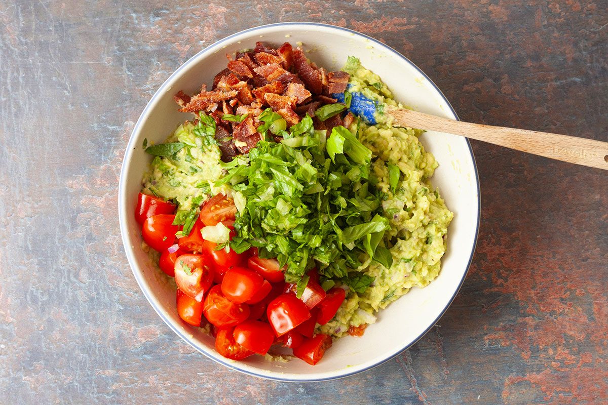 A white bowl filled with chopped lettuce, diced tomatoes, crumbled bacon, and mashed avocado, with a wooden spoon resting on the side. The bowl sits on a textured brown surface.
