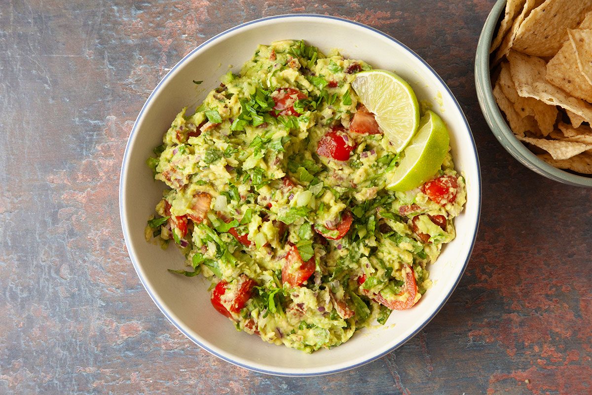 A bowl of guacamole with chopped tomatoes, onions, cilantro, and two lime wedges, next to a bowl of tortilla chips on a rustic surface.