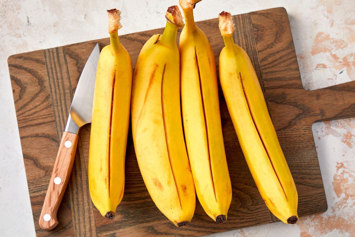 Overhead shot of cut banana peel lengthwise about 1/2 inch deep on wooden cutting board with a knife; leaving 1/2 inch at both ends; Open peel wider to form a pocket; all set on a marble surface;