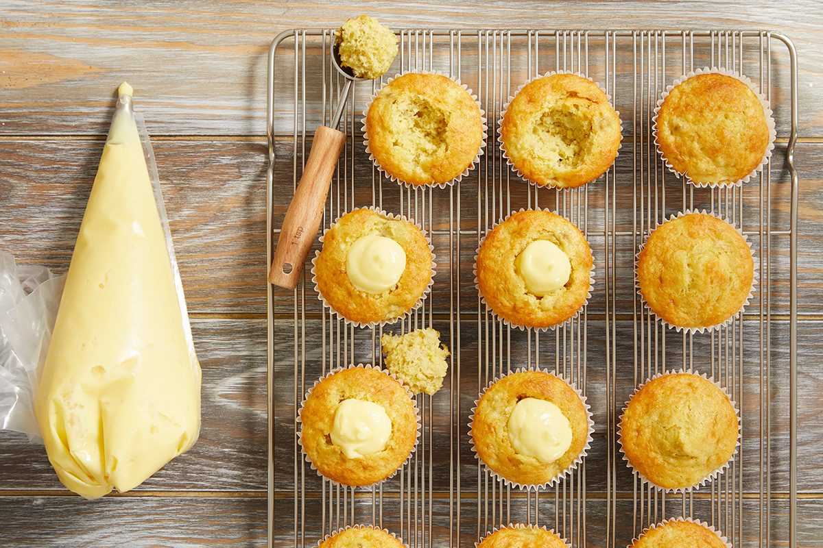 A cooling rack holds cupcakes with holes cut from their centers, some filled with cream. A melon baller tool and cupcake tops are nearby. A piping bag of yellow cream sits on the left side of a wooden surface.