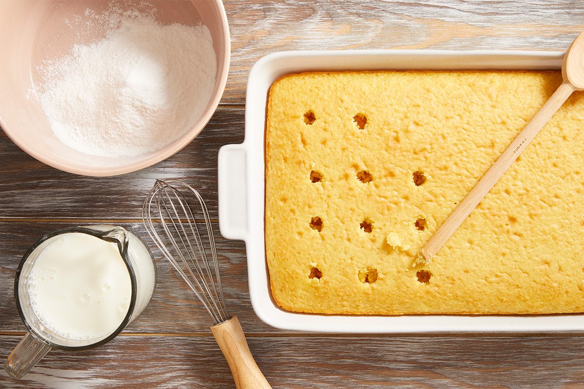 A rectangular baked cake in a white dish with holes poked in the top, next to a wooden spoon, a whisk, a bowl of powdered sugar, and a glass measuring cup of milk on a wooden surface.