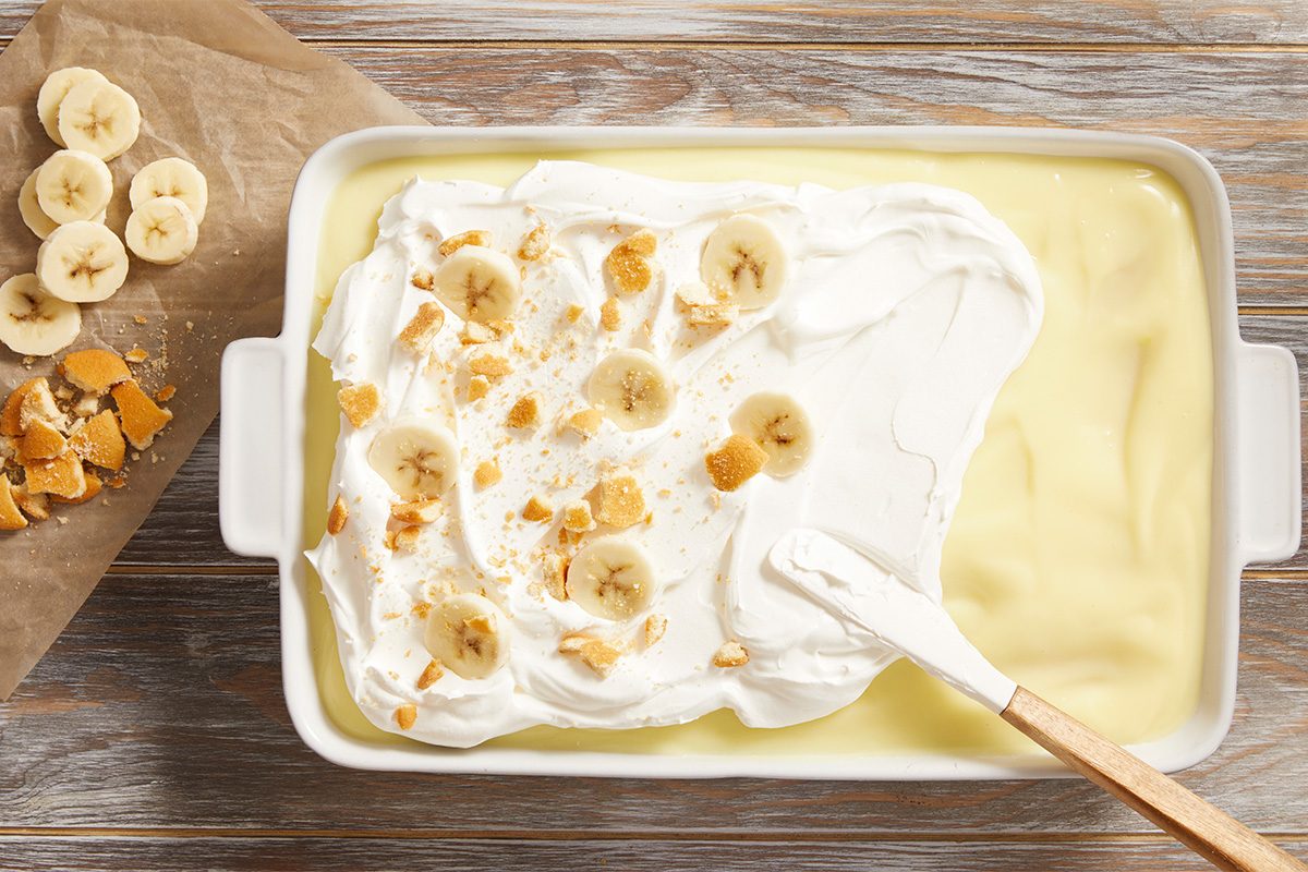 A baking dish filled with banana pudding, partially covered in whipped cream, banana slices, and crushed wafers. A spatula is spreading the topping. Sliced bananas and wafer pieces are on the side on parchment paper.