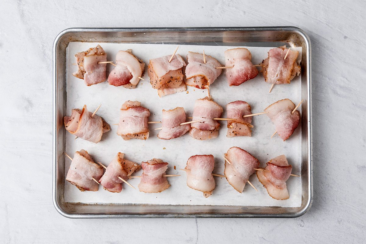 A baking tray lined with parchment paper holds rows of bite-sized pieces of food wrapped in bacon and secured with toothpicks, ready to be cooked.