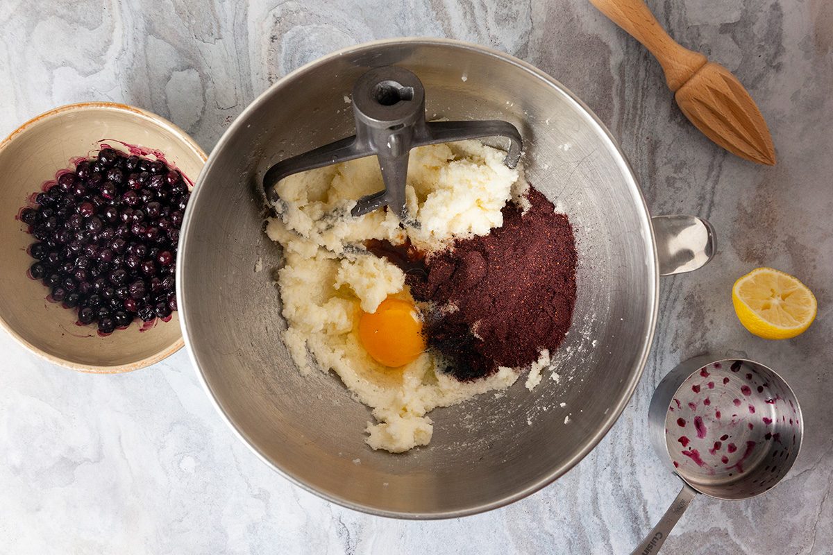 Overhead shot of all ingredients; in a small microwave safe bowl; microwave frozen blueberries on high 30 to 60 seconds or until slightly soft; set aside; place softened butter and sugar in the bowl of a stand mixer fitted with a paddle attachment; cream on medium to high speed until fluffy, 2 to 3 minutes, scraping the sides of the bowl as needed; beat in egg; vanilla extract; lemon juice and ground freeze dried blueberries until a soft dough forms; 1 to 2 minutes; wooden lemon squeezer is visible nearby; all set on a marble surface;