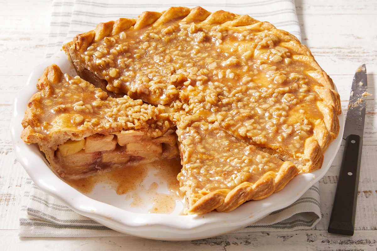 A caramel apple pie with a crumbly topping sits in a white pie dish. One slice has been removed, revealing the apple filling inside. A knife rests nearby on a striped cloth.