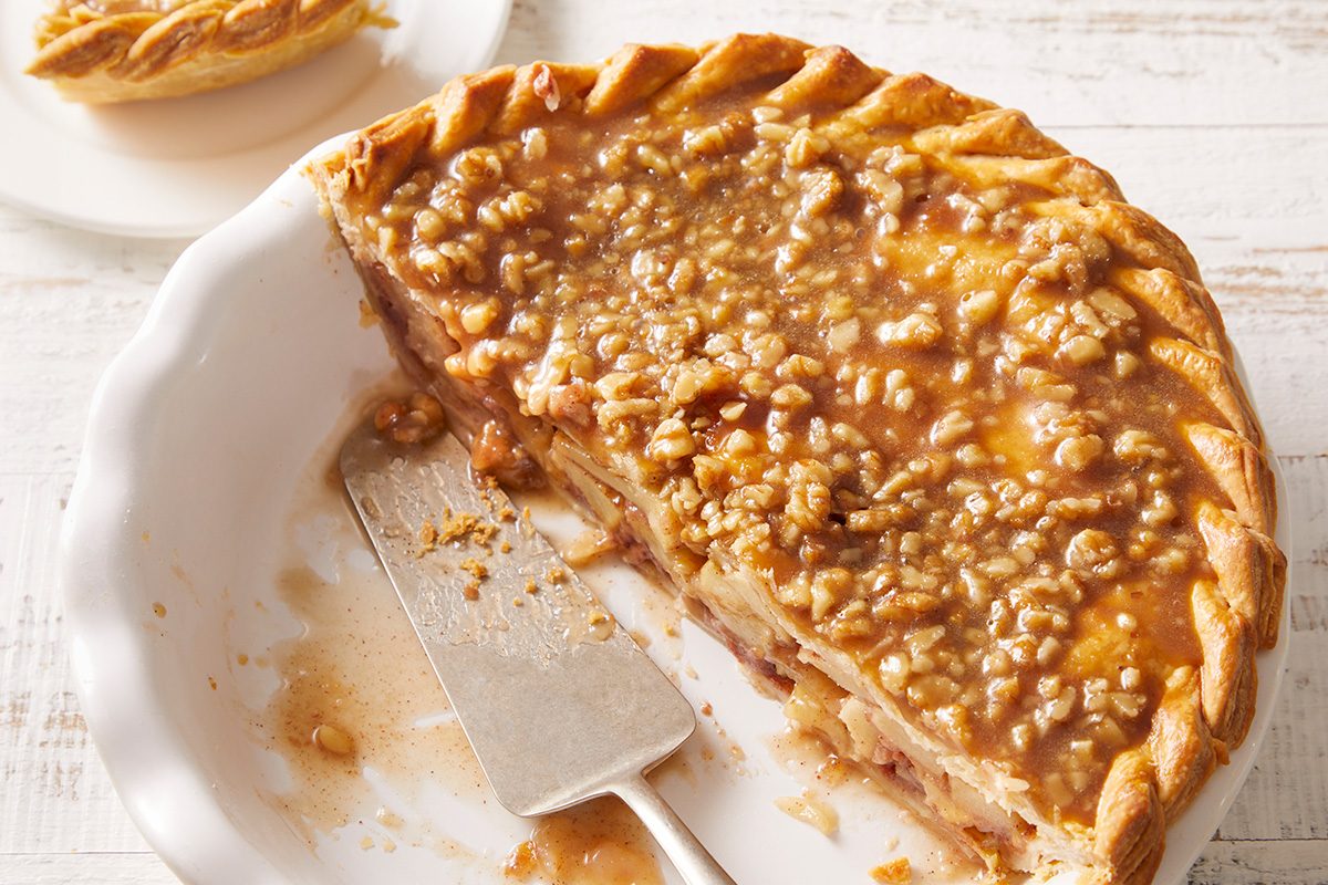 A partially sliced pie with a golden, crumbly topping sits in a white dish. A pie server rests inside, and a slice of pie is on a small plate in the background. The scene is set on a light wooden surface.