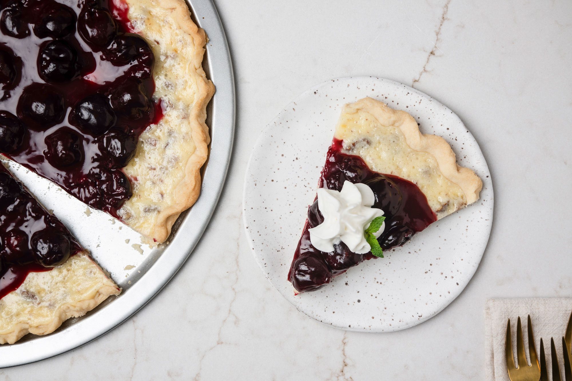 Overhead shot of Cherry Cheese Pizza on a pie plate; cut a small slice and served on small ceramic plate; Top with whipped cream and mint; forks are resting on a cream napkin nearby; all set on a white texture marble surface