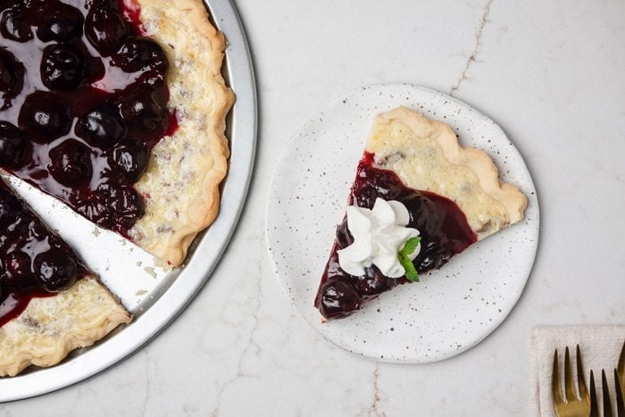 Overhead shot of Cherry Cheese Pizza on a pie plate; cut a small slice and served on small ceramic plate; Top with whipped cream and mint; forks are resting on a cream napkin nearby; all set on a white texture marble surface