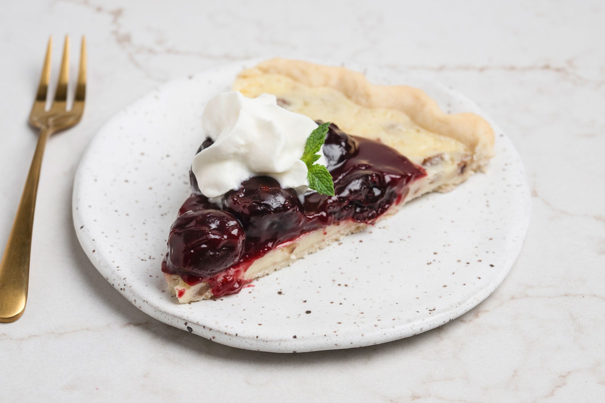 Closeup shot of Cherry Cheese Pizza; cut a small slice and served on small ceramic plate; Top with whipped cream and mint with a fork nearby; all set on a white texture marble surface