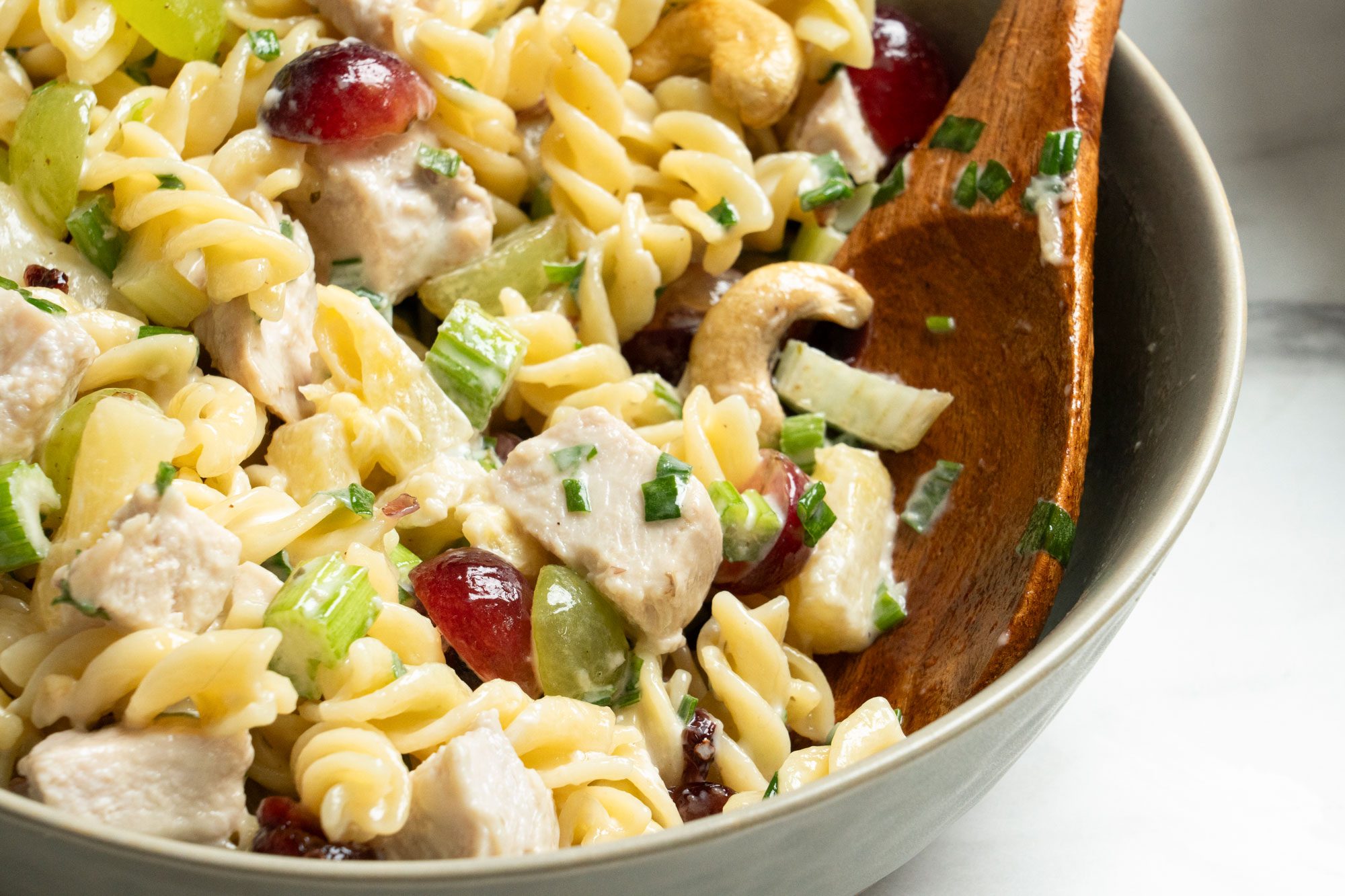 Closeup shot of Cashew-Chicken Rotini Salad served in a large bowl; with a wooden spoon; all set on a marble surface