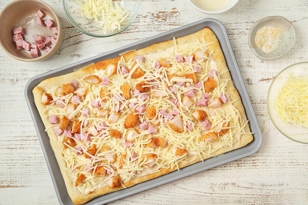A rectangular pizza dough on a baking sheet topped with shredded cheese, diced ham, and chicken pieces. Surrounding the tray are bowls with more cheese, ham cubes, and sauce on a white wooden surface.