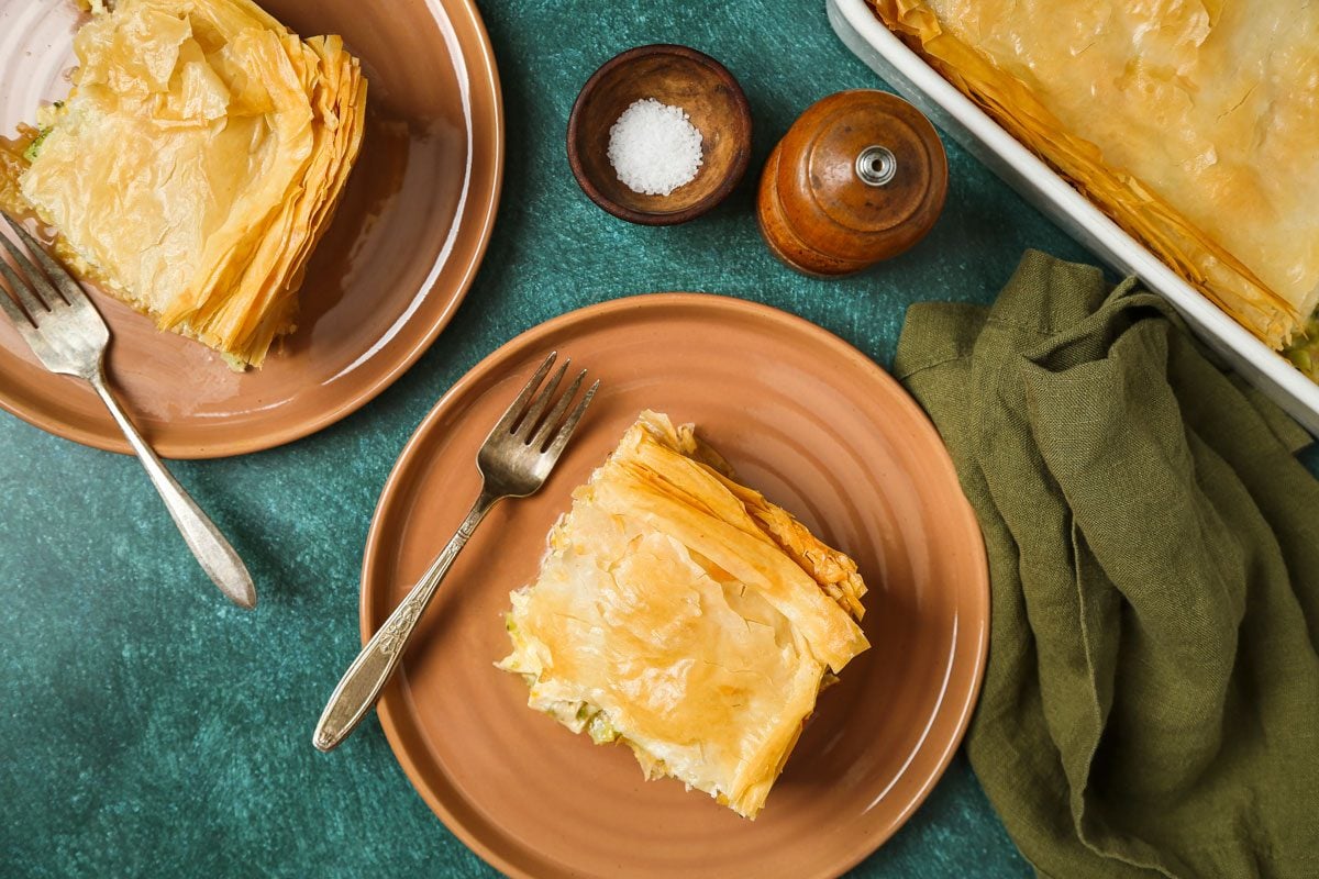 Two slices of golden; flaky pie rest on brown plates with forks; a baking dish holds more pie nearby; accompanied by a pepper grinder; salt bowl; and green napkin on a teal surface, creating a cozy, inviting scene