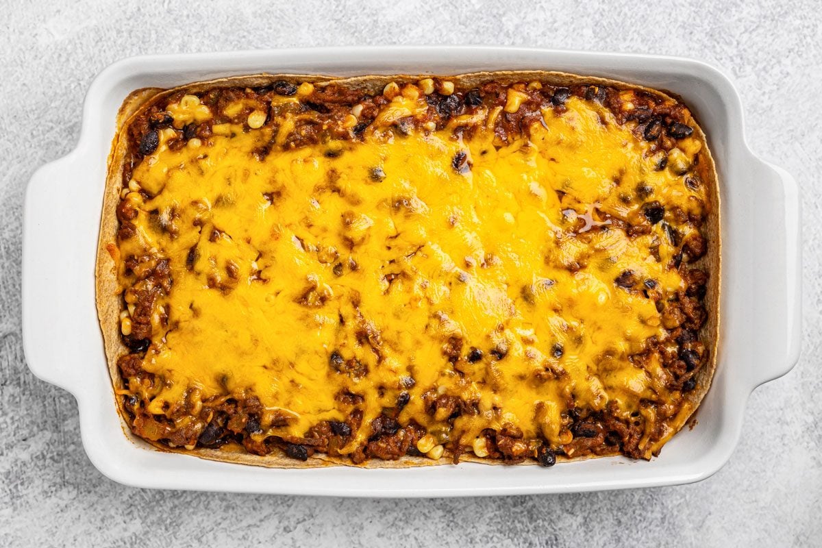overhead shot of a white rectangular baking dish filled with a baked casserole topped with melted cheddar cheese, ground meat, and beans, set on a light gray textured surface
