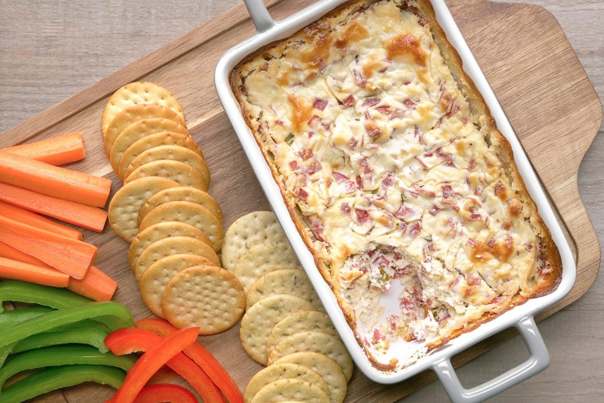 overhead shot of a rectangular dish of creamy baked dip with chopped ingredients sits on a wooden board, surrounded by round crackers, carrot sticks, and slices of green and red bell peppers