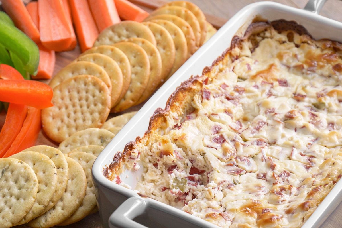 3/4th shot of a rectangular dish of creamy baked dip with chopped ingredients sits on a wooden board, surrounded by round crackers, carrot sticks, and slices of green and red bell pepper