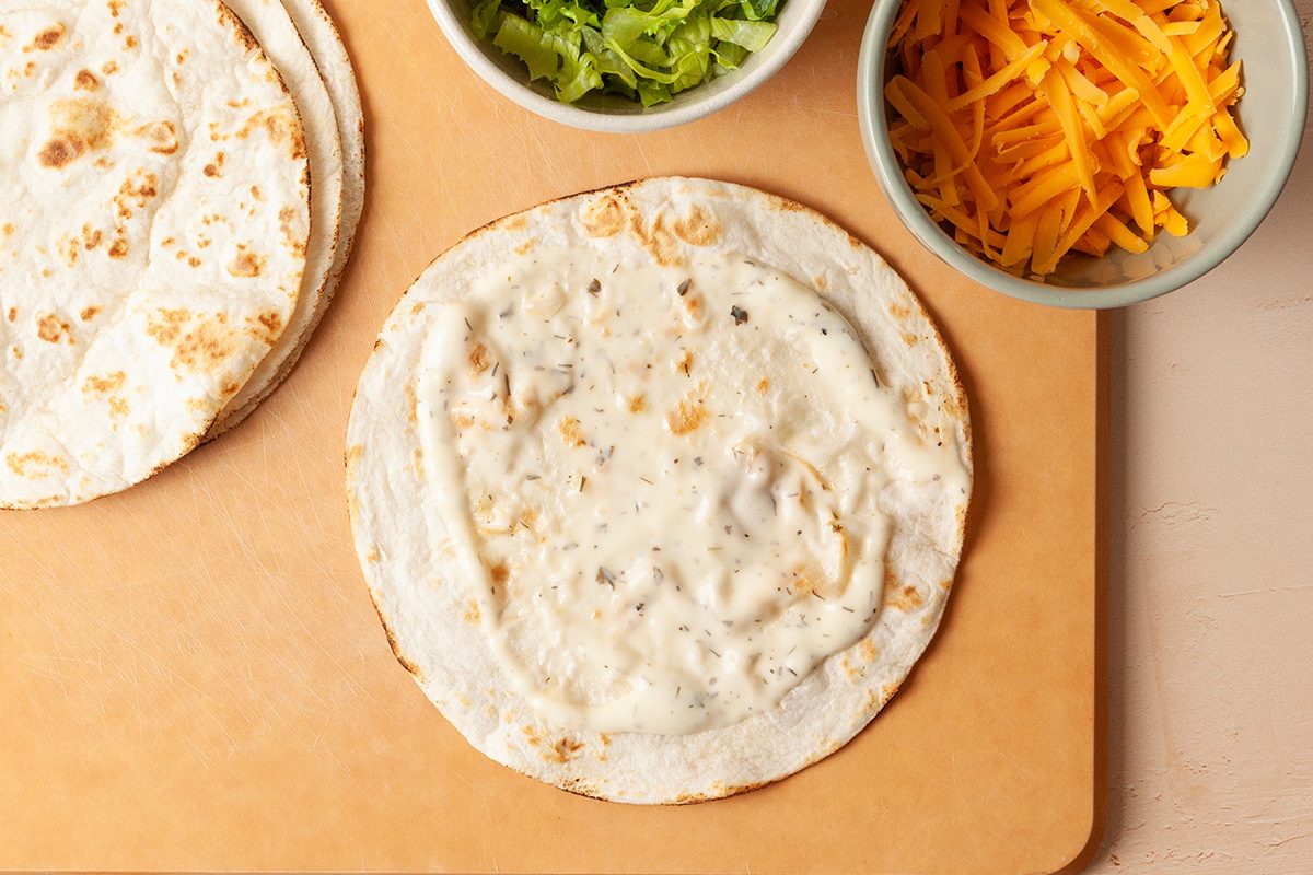 A cutting board with stacked flour tortillas, a bowl of shredded lettuce, a bowl of shredded cheddar cheese, and a plate with breaded chicken tenders on a light-colored surface.