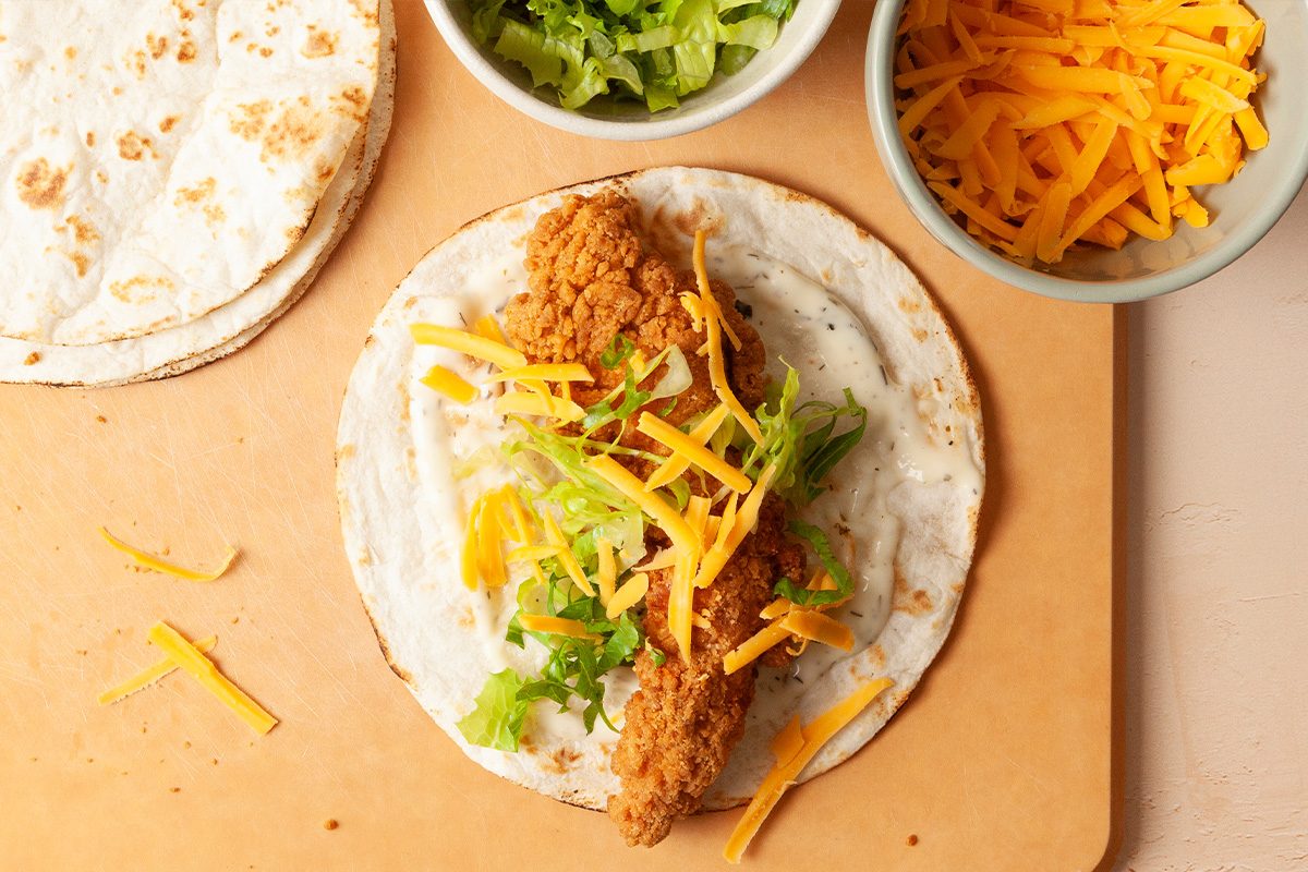 A soft tortilla with a crispy fried chicken strip, shredded lettuce, and cheddar cheese sits on a cutting board, with bowls of more shredded lettuce and cheese nearby.
