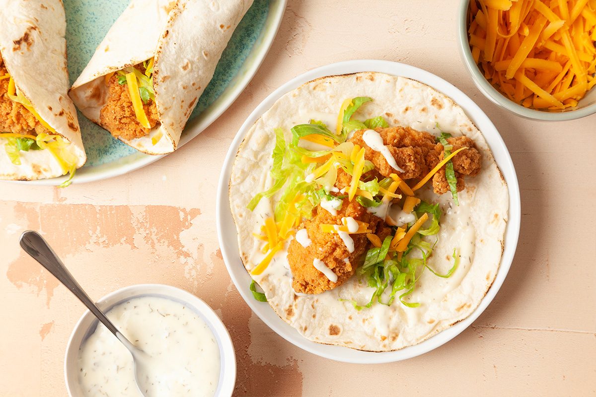 A flour tortilla topped with fried chicken, shredded lettuce, cheddar cheese, and a drizzle of white sauce on a plate, with more prepared wraps, shredded cheese, and sauce nearby.