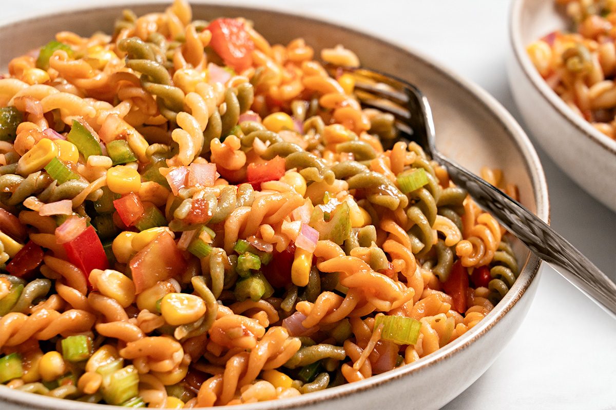 A close-up of a bowl of tri-color rotini pasta salad mixed with diced vegetables such as tomatoes, green peppers, red onions, and corn, with a fork resting in the bowl.