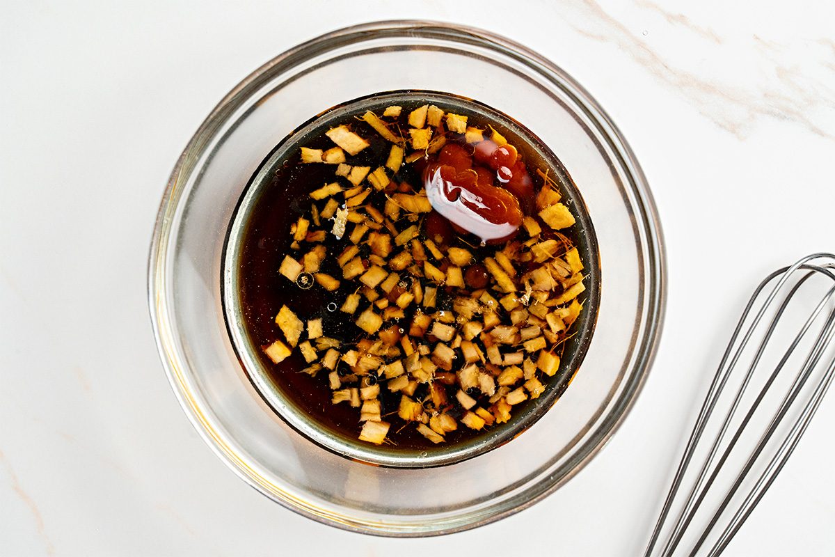 A glass bowl filled with a dark sauce containing chopped garlic or ginger and a dollop of ketchup, sitting on a white surface next to a metal whisk.