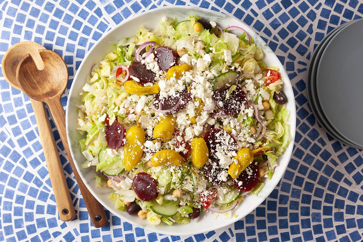 A bowl of fresh salad topped with feta cheese, cherry tomatoes, sliced red onions, olives, and beets sits on a blue and white mosaic table next to wooden serving utensils and stacked gray plates.