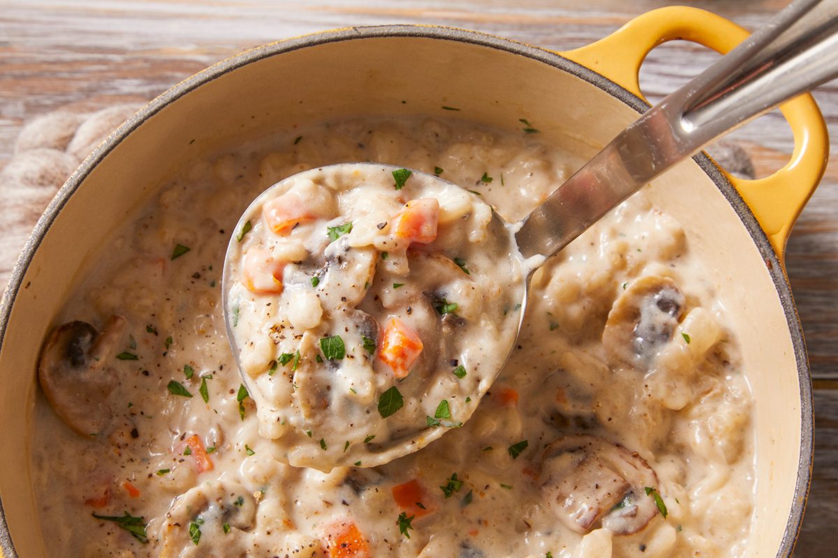 A close-up of a creamy vegetable stew in a yellow pot, with a ladle lifting a portion containing carrots, mushrooms, potatoes, and sprinkled herbs.