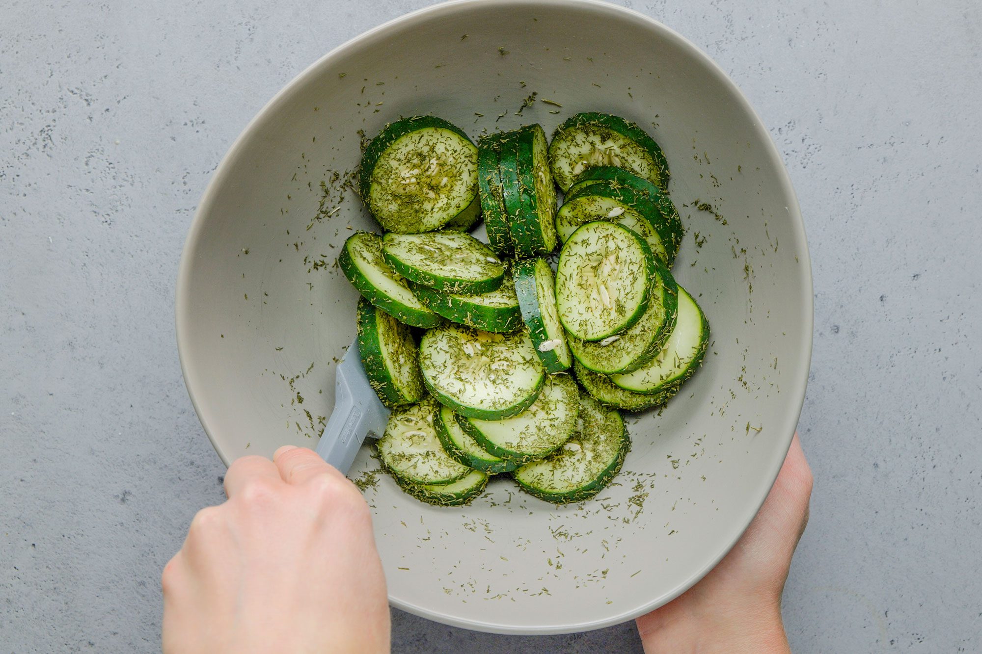 Overhead shot of another bowl with cucumber and dill being tossed; all set on a grey surface