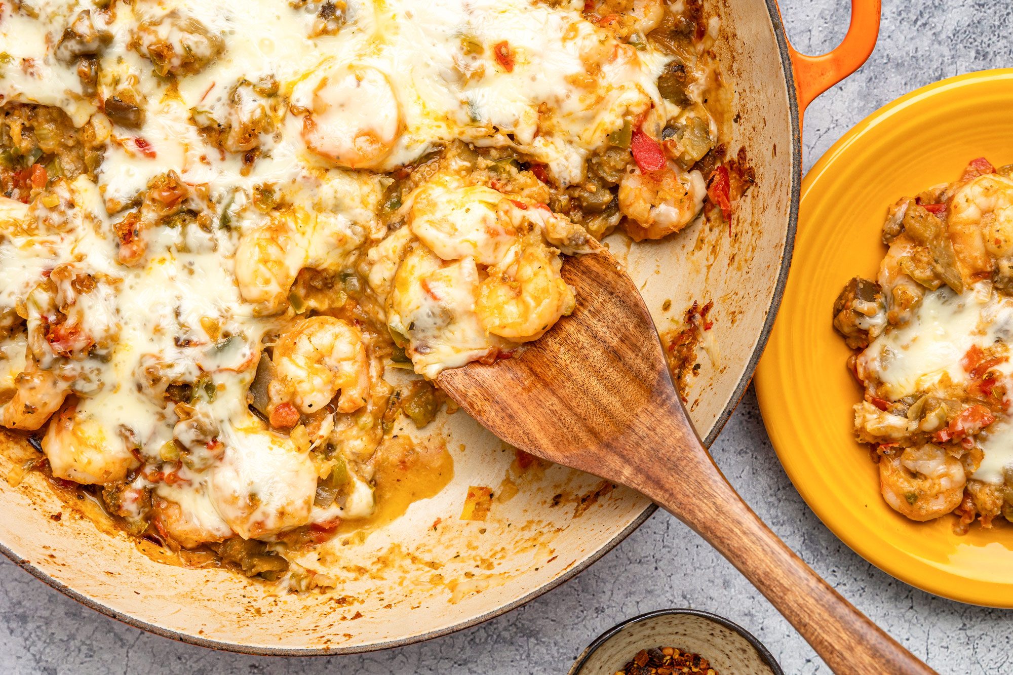 overhead shot of a skillet filled with cheesy shrimp and vegetables in a creamy sauce, with a wooden spoon serving a portion onto a yellow plate nearby